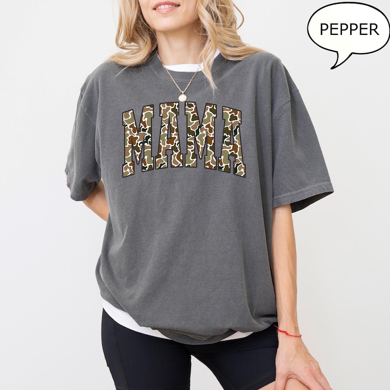 May include: A gray oversized t-shirt with a camo print "MAMA" graphic. The model is wearing black shorts and a silver necklace.