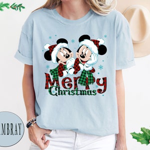 May include: Light blue t-shirt with a Christmas-themed design featuring Mickey Mouse and Minnie Mouse wearing Santa hats and scarves. The text "Merry Christmas" is printed below the characters.