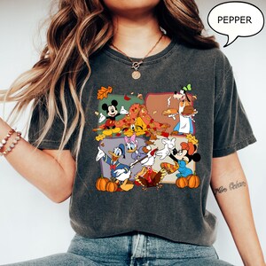 May include: Dark gray t-shirt featuring a colorful graphic of Disney characters celebrating autumn. Mickey Mouse, Minnie Mouse, Donald Duck, Daisy Duck, and Goofy are depicted with pumpkins and fall leaves. The word "PEPPER" is in a speech bubble.
