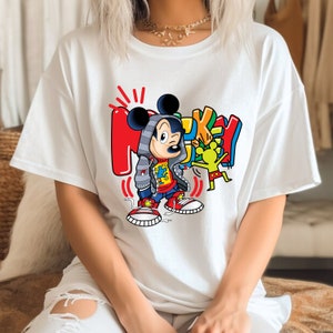 Pode incluir: Camiseta branca com um gr&aacute;fico colorido do Mickey Mouse vestindo um moletom cinza e sapatos vermelhos. O gr&aacute;fico tamb&eacute;m inclui a palavra "Mickey" em uma fonte colorida de estilo graffiti.