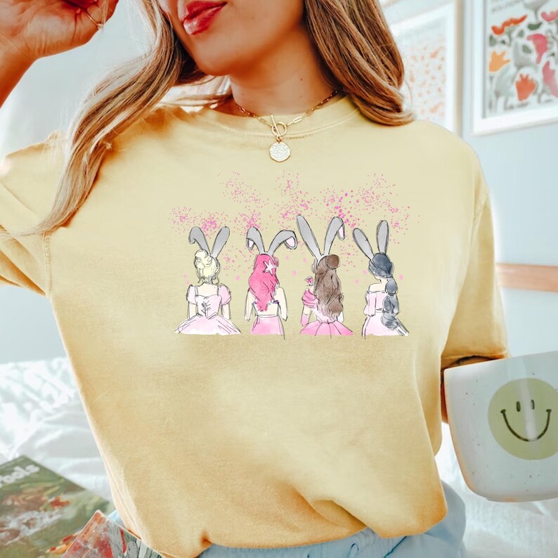 Puede incluir: Una camiseta amarilla con un gr&aacute;fico de cuatro mujeres con orejas de conejo. Las mujeres llevan vestidos rosas y tienen diferentes colores de pelo.