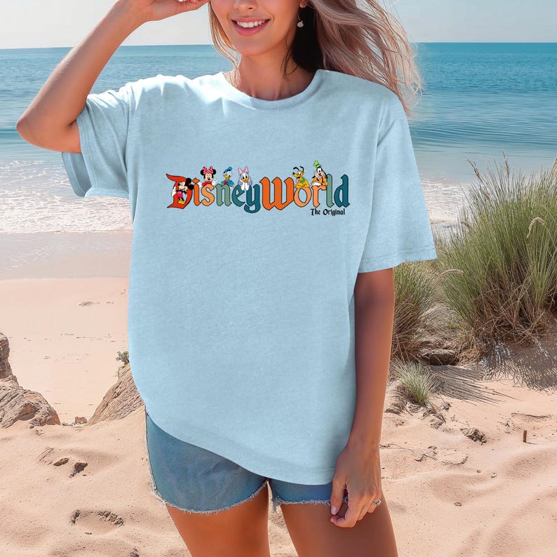 K&ouml;nnte beinhalten: Ein hellblaues T-Shirt mit einem Aufdruck von Disney-Figuren und dem Text "Disney World The Original".