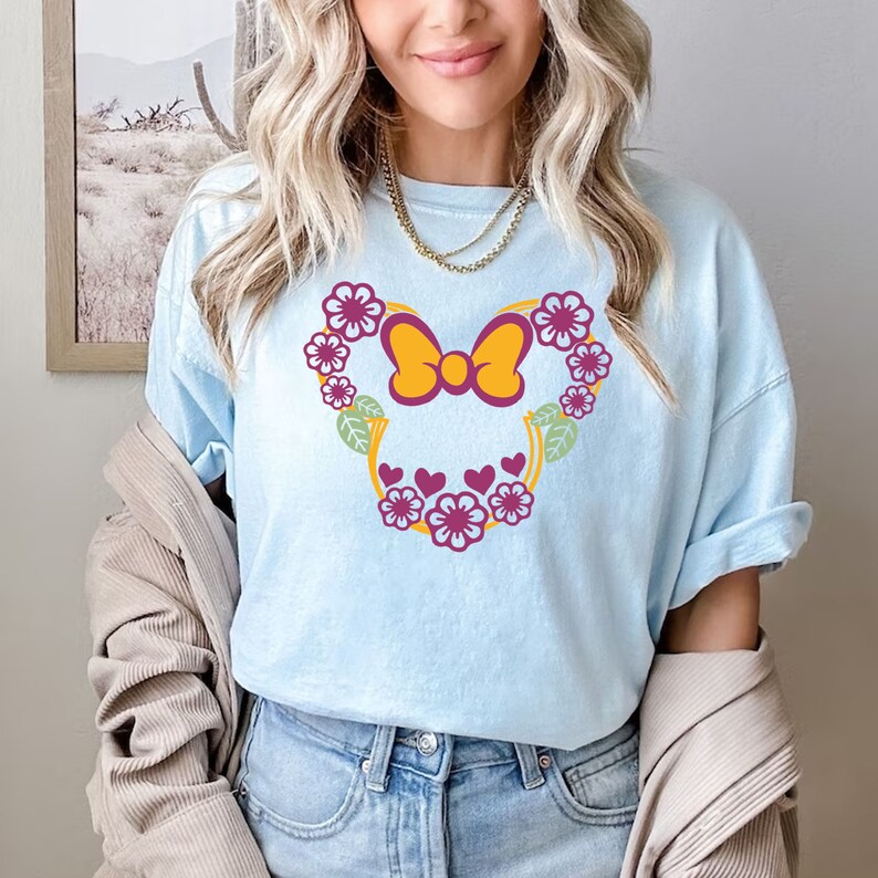 May include: Light blue t-shirt with a floral design in the shape of Minnie Mouse's head. The flowers are pink, yellow, and green. The bow is yellow with a purple center.