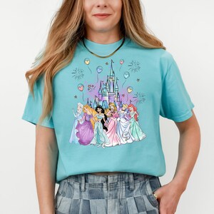 Puede incluir: Una camiseta azul turquesa con un gr&aacute;fico de las princesas de Disney frente al castillo de Cenicienta. Las princesas llevan sus vestidos caracter&iacute;sticos y est&aacute;n rodeadas de fuegos artificiales y globos.