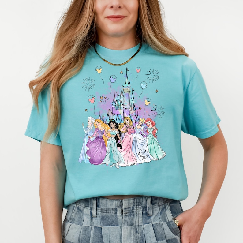 Puede incluir: Una camiseta azul turquesa con un gr&aacute;fico de las princesas de Disney frente al castillo de Cenicienta. Las princesas llevan sus vestidos caracter&iacute;sticos y est&aacute;n rodeadas de fuegos artificiales y globos.