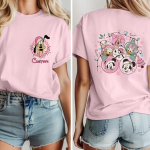 K&ouml;nnte beinhalten: Hellrosa T-Shirt mit einem Cartoon von Pluto mit Weihnachtsm&uuml;tze und Lichterketten, darunter das Wort "Custom". Auf der R&uuml;ckseite des Shirts sind Mickey, Minnie, Donald und Daisy in Weihnachtskugeln abgebildet.
