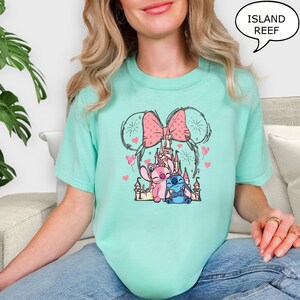 Puede incluir: Una camiseta turquesa clara con un gr&aacute;fico de un castillo, un lazo rosa y orejas de rat&oacute;n. El gr&aacute;fico incluye los personajes Stitch y Angel, con corazones y fuegos artificiales. El texto "ISLAND REEF" est&aacute; en un bocadillo.