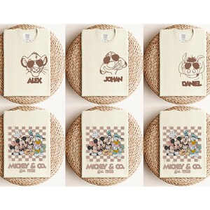 May include: Cream-colored t-shirts featuring cartoon characters with sunglasses. The shirts display characters like Simba, Timon, and Pumbaa, along with names. Other shirts showcase Mickey Mouse, Donald Duck, and Goofy with the text "MICKEY & CO. Est. 1928."