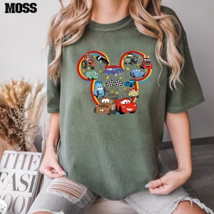 Puede incluir: Una camiseta verde con una silueta de Mickey Mouse formada por varios coches de dibujos animados de la pel&iacute;cula Cars de Disney. Los coches son de varios colores, incluyendo rojo, azul, amarillo y verde.