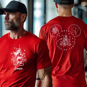 K&ouml;nnte beinhalten: Rotes T-Shirt mit einem wei&szlig;en Aufdruck einer Cartoon-Maus vor einem Schloss und dem Namen "James" darunter. Auf der R&uuml;ckseite befindet sich ein Schlossdesign in einer Mickey-Mouse-Kopfkontur mit dem Text "The Happiest Place on Earth".
