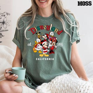May include: Green t-shirt with a vintage-style Disneyland logo featuring Mickey Mouse, Minnie Mouse, Donald Duck, Goofy, and Pluto in Christmas attire. The text reads "Disneyland est. 1955 CALIFORNIA".