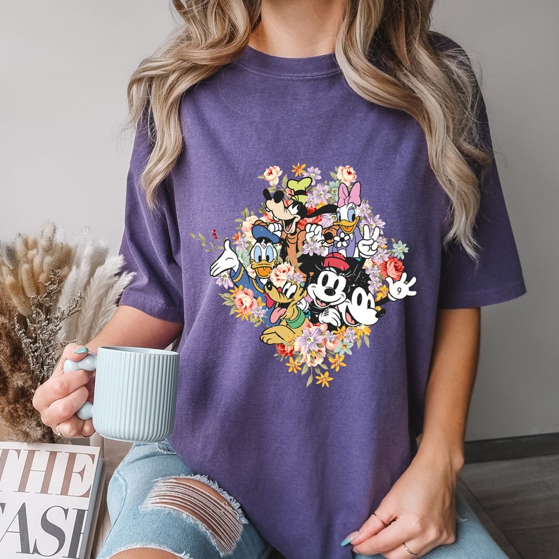 May include: Purple t-shirt featuring a graphic print of Mickey Mouse and his friends surrounded by flowers.