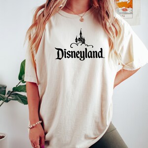 Puede incluir: Camiseta color crema con la palabra "Disneyland" en fuente negra estilizada, con un gr&aacute;fico de castillo encima. La camiseta tiene mangas cortas y un corte holgado, adecuada para uso casual.