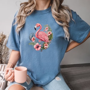 K&ouml;nnte beinhalten: Ein blaues T-Shirt mit einem pinkfarbenen Flamingo und einem Blumendesign. Der Flamingo steht auf einem Bein und hat den Kopf zur Seite geneigt. Die Blumen sind pink, wei&szlig; und gr&uuml;n.