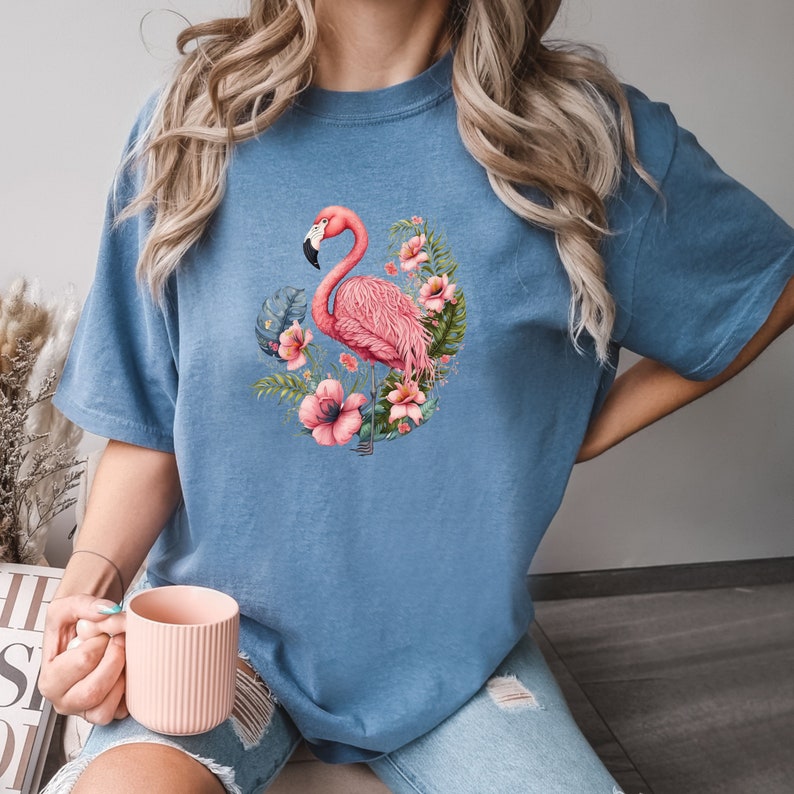 K&ouml;nnte beinhalten: Ein blaues T-Shirt mit einem pinkfarbenen Flamingo und einem Blumendesign. Der Flamingo steht auf einem Bein und hat den Kopf zur Seite geneigt. Die Blumen sind pink, wei&szlig; und gr&uuml;n.