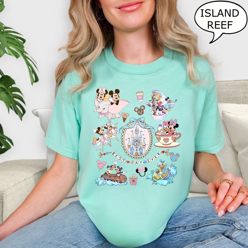 Puede incluir: Camiseta de color verde azulado claro con un gr&aacute;fico colorido de personajes de dibujos animados, incluyendo Mickey y Minnie Mouse, con el texto "BEST DAY EVER". La camiseta tambi&eacute;n tiene el texto "ISLAND REEF" en un bocadillo.