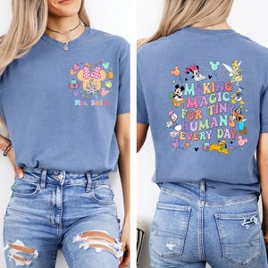 May include: A blue short-sleeved t-shirt with a colourful graphic on the front and back. The front features a Minnie Mouse design with a castle and the name "Mrs. Smi". The back says "Making Magic for Tiny Humans Every Day" with Disney characters.