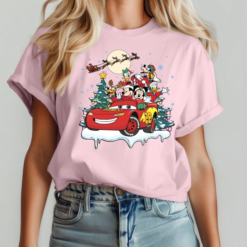 May include: Pink t-shirt with a Christmas-themed graphic featuring Mickey Mouse, Donald Duck, Goofy, and Pluto riding in a red Lightning McQueen car. The characters are wearing Santa hats and the car is decorated with Christmas lights.
