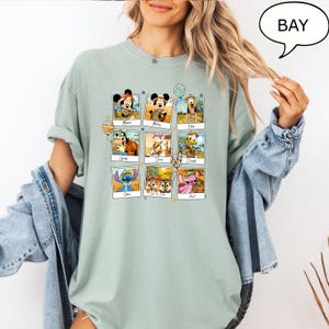 Puede incluir: Una camiseta verde claro con un gr&aacute;fico de una cuadr&iacute;cula de nueve personajes de dibujos animados del Libro de la Selva de Disney. Cada personaje est&aacute; en un marco cuadrado con su nombre debajo. Los personajes son: Mowgli, Mickey, Pluto, Goofy, Dory, Donald, Stitch, Simba & Nala y Angel.