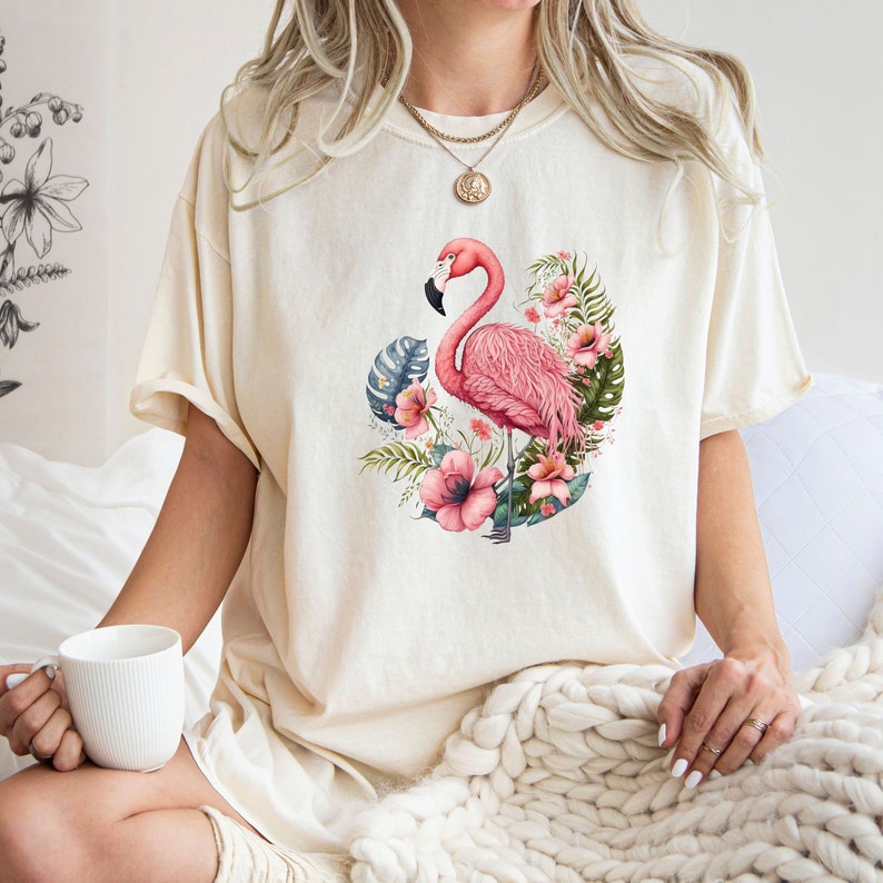 K&ouml;nnte beinhalten: Ein wei&szlig;es T-Shirt mit einem pinkfarbenen Flamingo und tropischen Blumen, die auf der Vorderseite aufgedruckt sind.
