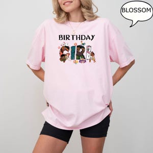 May include: Pink t-shirt with the text "Birthday Girl" in black letters. The letters are decorated with colourful cartoon characters.