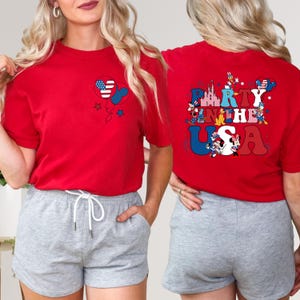 K&ouml;nnte beinhalten: Rotes T-Shirt mit einem Mickey-Mouse-f&ouml;rmigen Ballon mit einem Design der amerikanischen Flagge. Auf der R&uuml;ckseite des Shirts steht der Text "Party in the USA" mit patriotischen Bildern. Das Shirt wird mit grauen Shorts getragen.