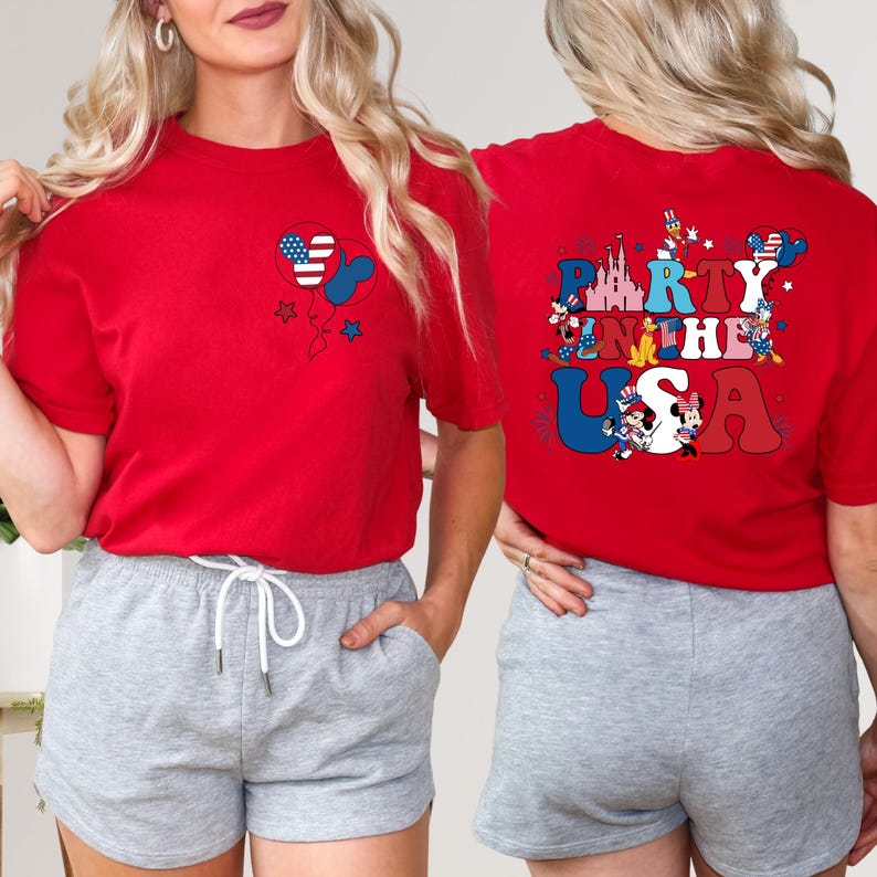 K&ouml;nnte beinhalten: Rotes T-Shirt mit einem Mickey-Mouse-f&ouml;rmigen Ballon mit einem Design der amerikanischen Flagge. Auf der R&uuml;ckseite des Shirts steht der Text "Party in the USA" mit patriotischen Bildern. Das Shirt wird mit grauen Shorts getragen.