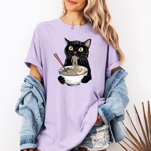 Puede incluir: Una camiseta morada con un gato negro comiendo fideos ramen de un taz&oacute;n blanco. El gato tiene ojos amarillos y lleva una diadema roja.