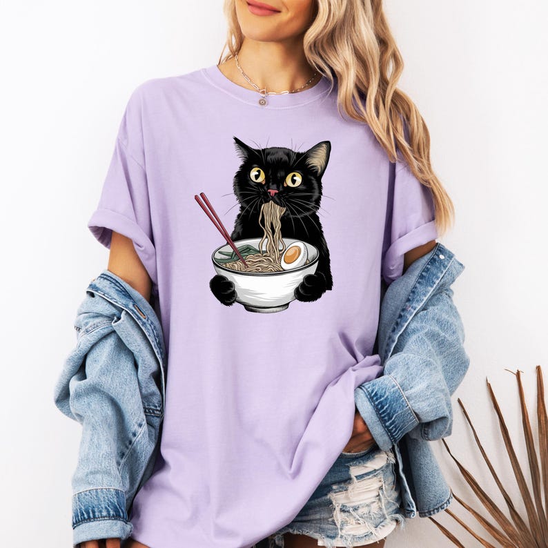 Puede incluir: Una camiseta morada con un gato negro comiendo fideos ramen de un taz&oacute;n blanco. El gato tiene ojos amarillos y lleva una diadema roja.