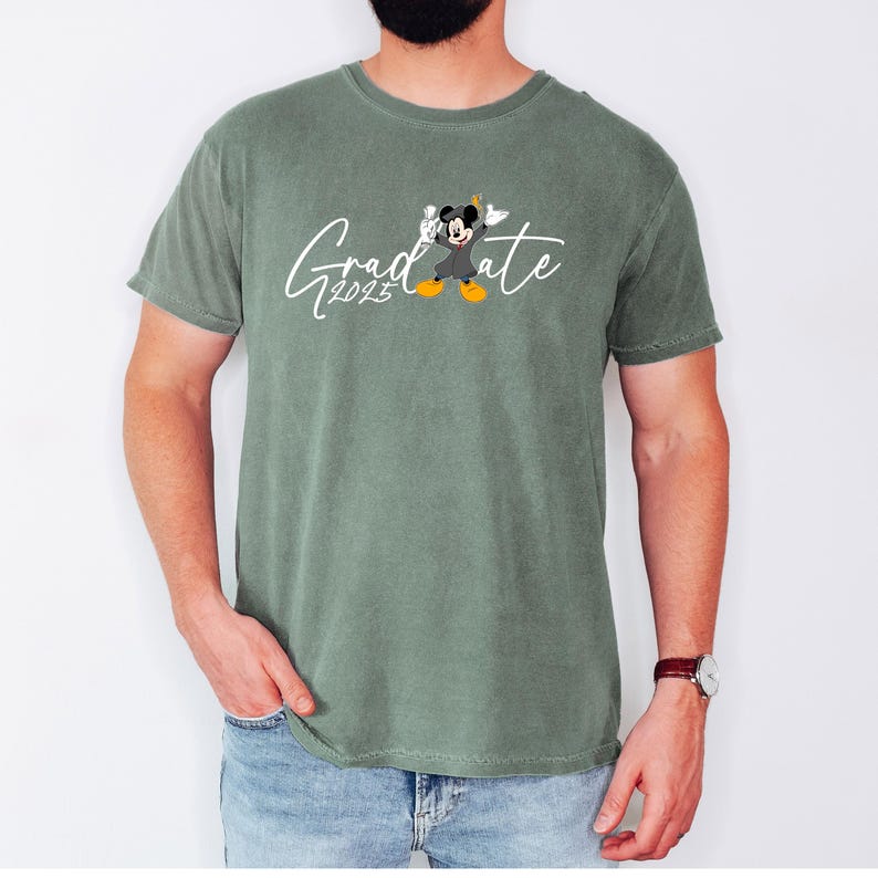 Puede incluir: Una camiseta verde con un gr&aacute;fico blanco de Mickey Mouse con un gorro de graduaci&oacute;n y una bata. El texto "Graduate 2023" est&aacute; escrito en cursiva blanca.