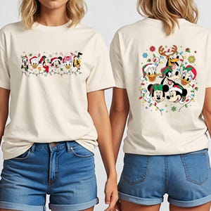May include: Off-white t-shirts featuring a festive Disney character design. The front of the shirt displays a line of characters with Christmas lights, while the back showcases a larger group of characters with holiday decorations.