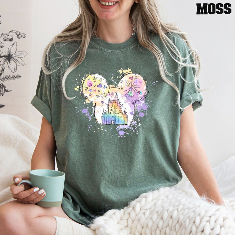 Puede incluir: Una camiseta verde con un gr&aacute;fico colorido de una cabeza de Mickey Mouse con el castillo de Cenicienta en su interior. El gr&aacute;fico est&aacute; rodeado de salpicaduras de pintura de colores.