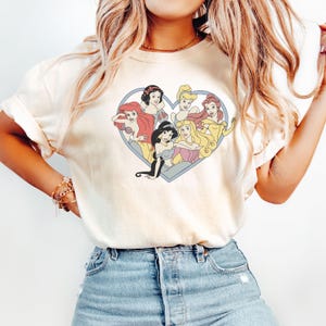 Pu&ograve; includere: Una maglietta bianca con una grafica di sette principesse Disney a forma di cuore. Le principesse sono Ariel, Belle, Cenerentola, Jasmine, Mulan, Biancaneve e Aurora.