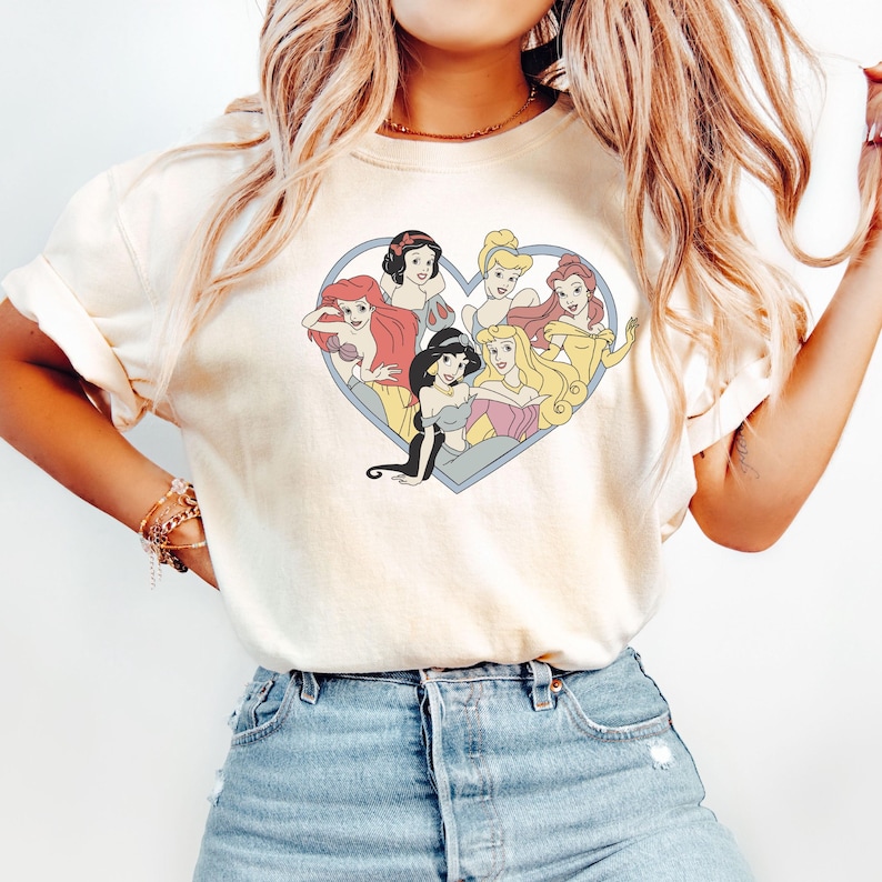 Pu&ograve; includere: Una maglietta bianca con una grafica di sette principesse Disney a forma di cuore. Le principesse sono Ariel, Belle, Cenerentola, Jasmine, Mulan, Biancaneve e Aurora.