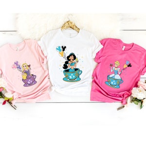 May include: Three white t-shirts with different Disney princesses in teacups. The princesses are Jasmine, Rapunzel, and Cinderella. Each princess is holding a balloon and a small object. The t-shirts are on a white wooden surface.