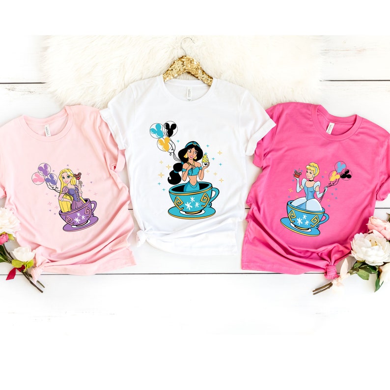 May include: Three white t-shirts with different Disney princesses in teacups. The princesses are Jasmine, Rapunzel, and Cinderella. Each princess is holding a balloon and a small object. The t-shirts are on a white wooden surface.