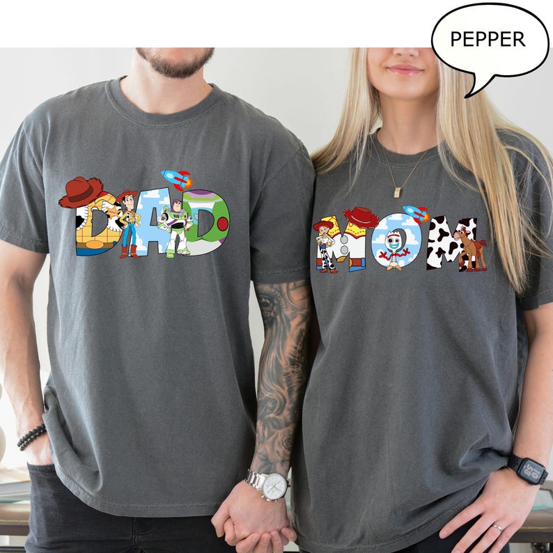 May include: Two grey t-shirts with the words "DAD" and "MOM" in colourful Toy Story-themed designs. The "DAD" shirt features Woody, Buzz Lightyear, and other characters. The "MOM" shirt has similar characters. The shirts are short-sleeved.