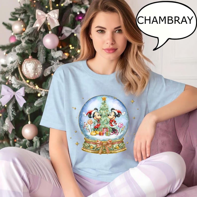 May include: Light blue t-shirt with a snow globe graphic featuring Mickey and Minnie Mouse in Christmas attire. The shirt has the word "CHAMBRAY" in a speech bubble. The person is wearing light purple and white striped pajama pants.