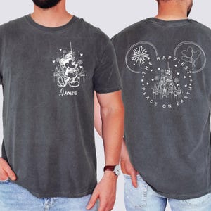 K&ouml;nnte beinhalten: Dunkelgraues T-Shirt mit wei&szlig;er Grafik von Mickey Mouse vor einem Schloss und Feuerwerk. Der Name "James" ist darunter gedruckt. Auf der R&uuml;ckseite befindet sich ein Mickey Mouse-Kopf-Design mit dem Text "The Happiest Place on Earth."