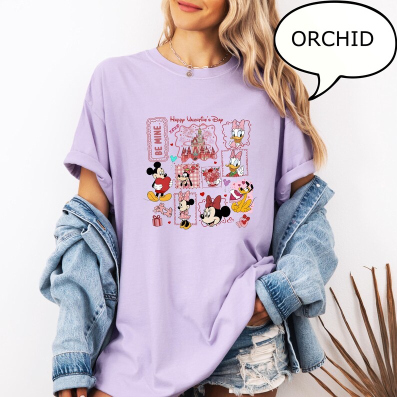 May include: A light purple t-shirt featuring a Valentine's Day graphic with Disney characters. The graphic includes Mickey and Minnie Mouse, Daisy Duck, and a castle. The shirt has rolled-up sleeves and the word "ORCHID" in a speech bubble.