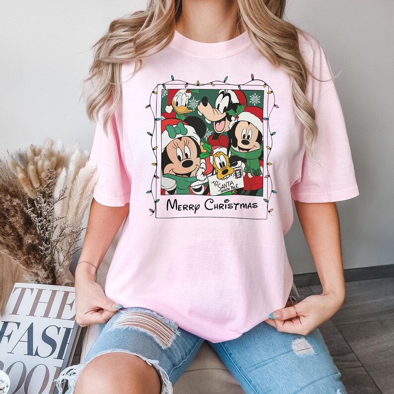 May include: Pink t-shirt with a Christmas-themed graphic featuring Mickey Mouse, Minnie Mouse, Donald Duck, and Goofy. The graphic is a vintage-style photo with a string of Christmas lights around it. The text "Merry Christmas" is printed below the graphic.