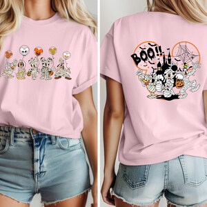 Puede incluir: Camiseta rosa claro con un dise&ntilde;o de Halloween. La parte delantera muestra personajes de dibujos animados momificados sosteniendo globos. La parte trasera tiene la palabra "Boo!!" y m&aacute;s personajes en un dise&ntilde;o circular. La camiseta es de material suave.