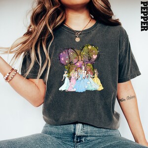 Puede incluir: Camiseta gris oscuro con un gr&aacute;fico de princesas de Disney y un castillo, dentro de un gran dise&ntilde;o de lazo. El lazo est&aacute; decorado con corazones y lunares. La palabra "PEPPER" est&aacute; impresa verticalmente en el lado derecho de la camiseta.