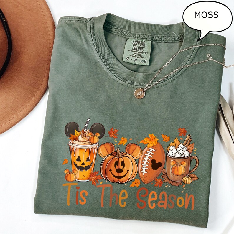 May include: A moss green Comfort Colors t-shirt with a fall-themed graphic. The graphic features a pumpkin spice latte with Mickey Mouse ears, a pumpkin with a Mickey Mouse face, a football with a Mickey Mouse head, and a mug of marshmallows. The words 'Tis The Season' are printed in orange and brown.