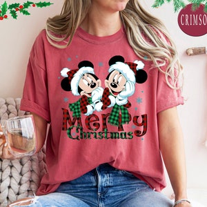 May include: Red t-shirt with a Christmas themed graphic featuring Mickey Mouse and Minnie Mouse wearing Santa hats. The text "Merry Christmas" is printed in black and red plaid.