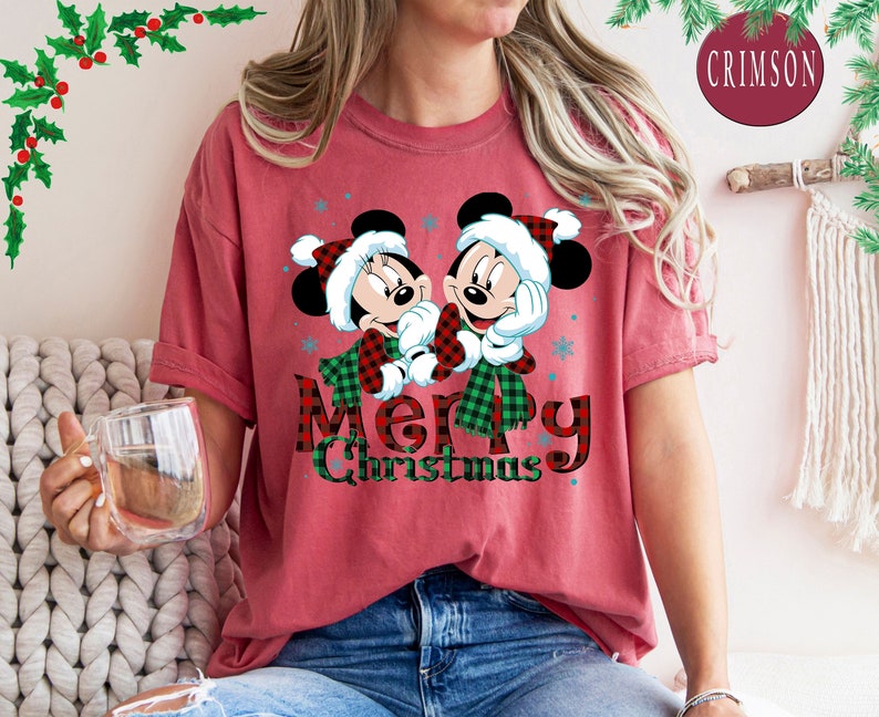 May include: Red t-shirt with a Christmas themed graphic featuring Mickey Mouse and Minnie Mouse wearing Santa hats. The text "Merry Christmas" is printed in black and red plaid.