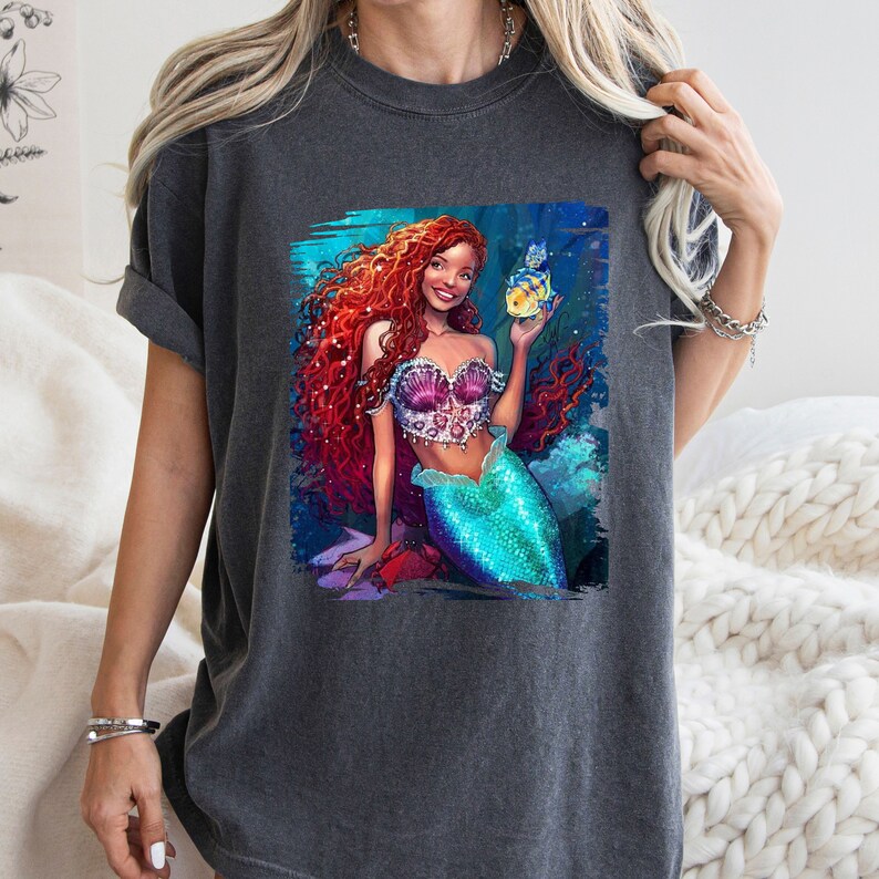 May include: A dark grey t-shirt featuring a colourful illustration of a mermaid with long red hair holding a yellow fish. The mermaid is wearing a seashell bra and a turquoise tail.