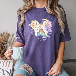 Puede incluir: Camiseta morada con un gr&aacute;fico colorido de un castillo dentro de una silueta de cabeza de Mickey Mouse. El gr&aacute;fico tiene un esquema de color arco&iacute;ris y un efecto acuarela.