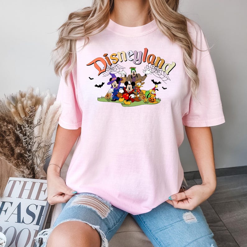 Puede incluir: Una camiseta rosa con un gr&aacute;fico de Mickey Mouse y sus amigos vestidos con disfraces de Halloween. El gr&aacute;fico incluye el texto "Disneyland" en una fuente colorida.