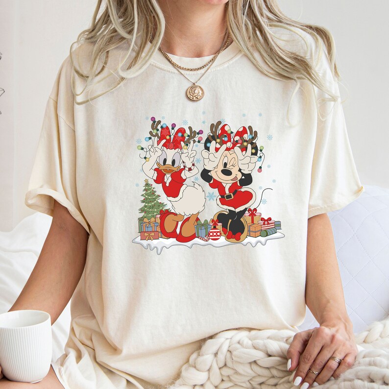 Puede incluir: Una camiseta blanca con un gr&aacute;fico navide&ntilde;o que presenta a Mickey Mouse y Daisy Duck con gorros de Pap&aacute; Noel y cuernos de reno. El gr&aacute;fico tambi&eacute;n incluye un &aacute;rbol de Navidad, regalos y copos de nieve.